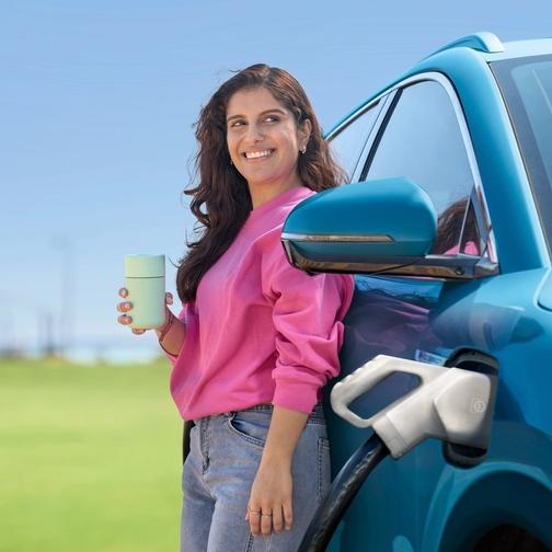 woman drinking from re-usable cup while charging electrical vehicle