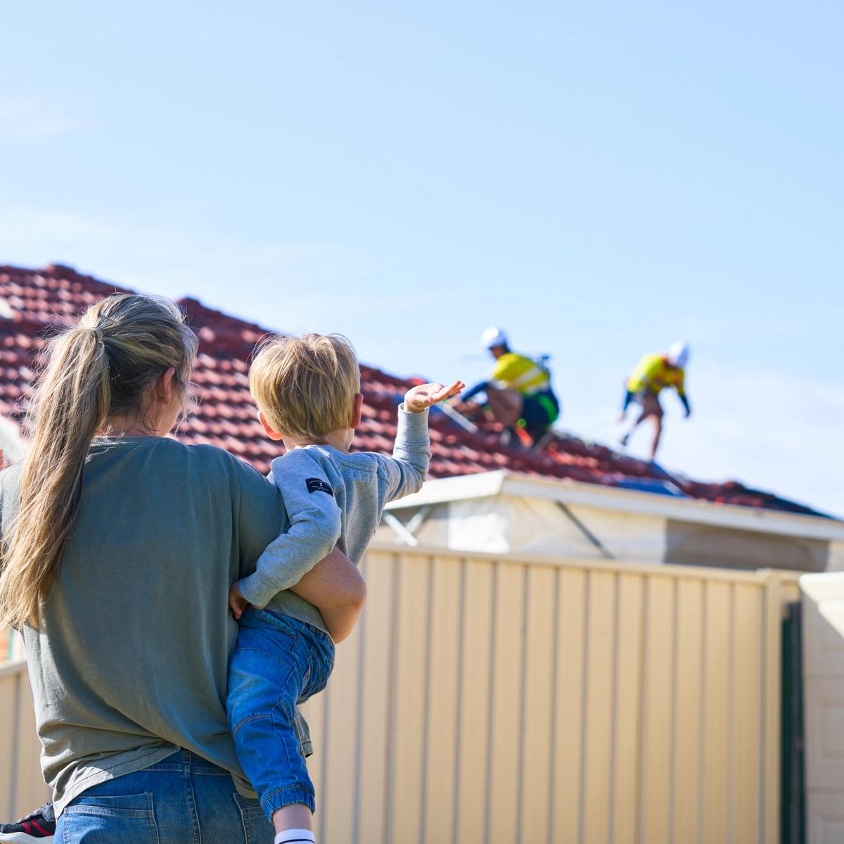 a mother holding her son and waving at workers on a roof installing solar panels